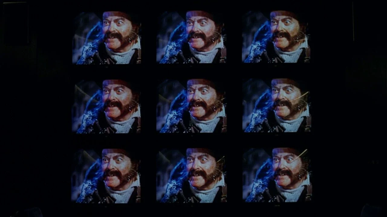 Scrooged (1988) movie still: wide — A wall displays a grid of nine identical monitors in a dark room. Each screen shows the same image of a man with…
