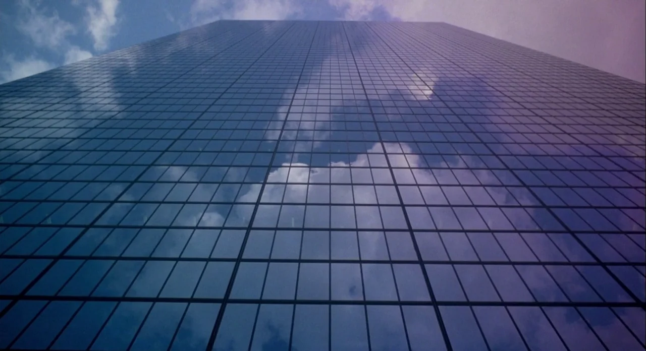 Koyaanasquatsi (1982) movie still: establishing — A tall modern skyscraper with a glass curtain wall dominates the frame. White clouds and blue sky…