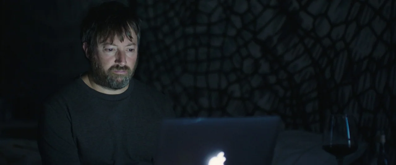 Greed (2019) movie still: medium — A dimly lit room features a patterned backdrop obscured by dark shadows. A bearded man looks at an open laptop…