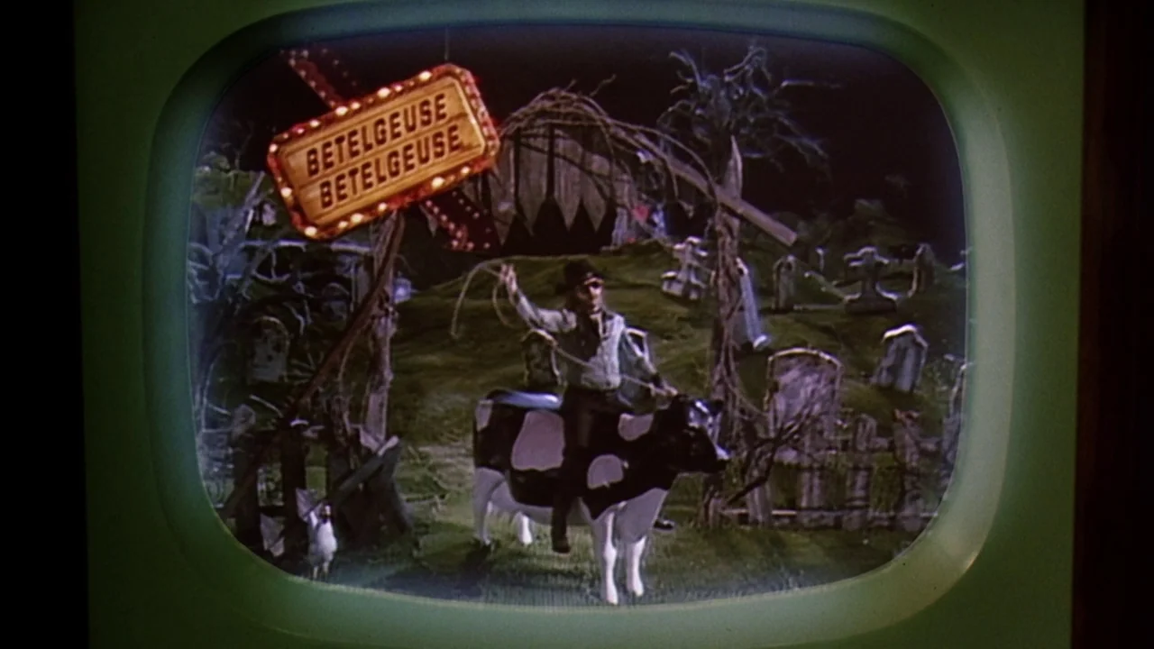 Beetlejuice (1988) movie still: medium — A miniature graveyard model features a stylized neon sign displaying the text Betelgeuse twice. A man sits…