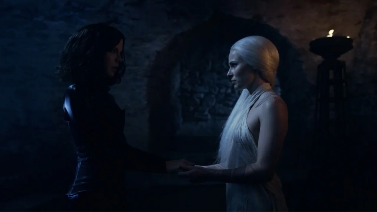 Underworld: Blood Wars (2016) movie still: medium — A dimly lit room with rough stone walls and a distant fire pit fixture. Two women hold hands…