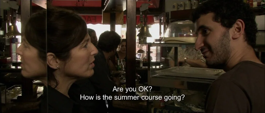 Genova (2008) movie still: medium — An interior cafe setting features a wooden counter and a glass display case. A woman and a man engage in…