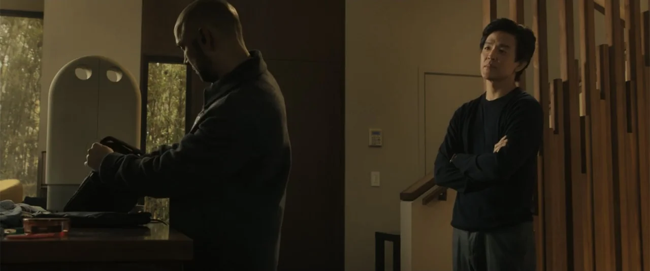 Afraid (2024) movie still: medium — A modern interior with vertical wooden slats on the right and a window background. Two men are standing near a…