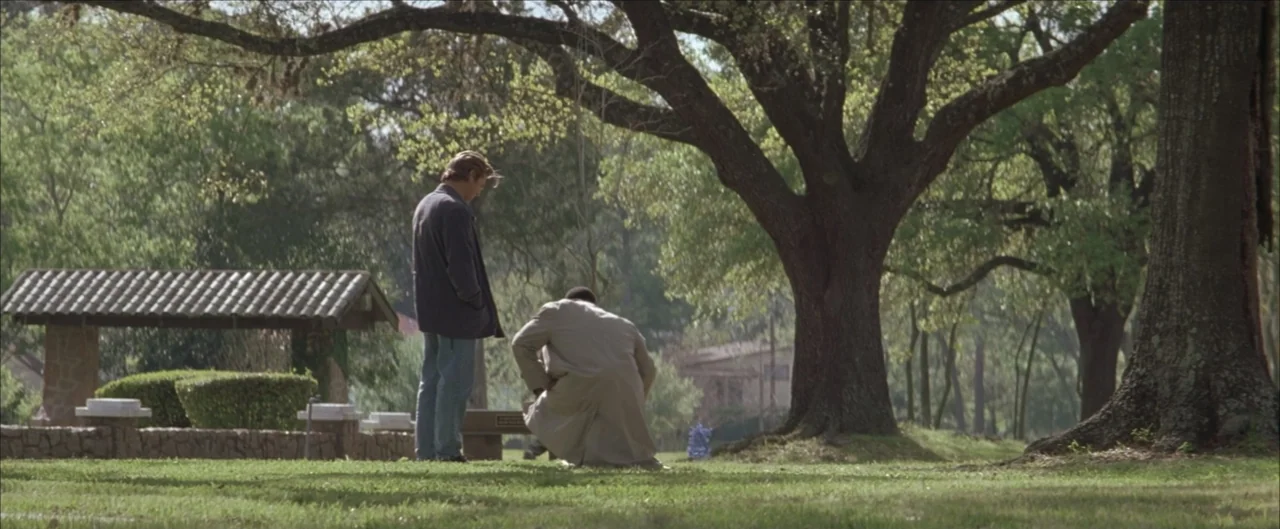 Arlington Road (1999) movie still: wide — A quiet cemetery with large oak trees and scattered grave markers under daylight. One man stands over a…