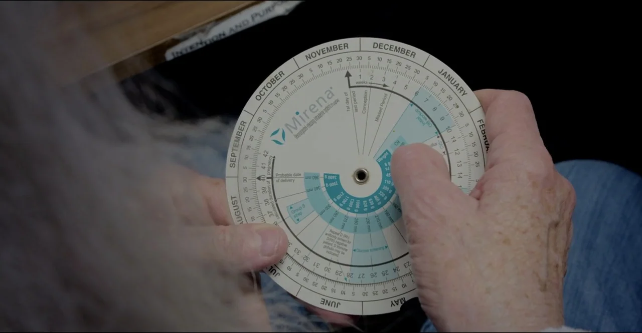 The Devil Is Busy (2024) movie still: closeup — An interior space features two hands holding a paper circular medical pregnancy calculator wheel.…