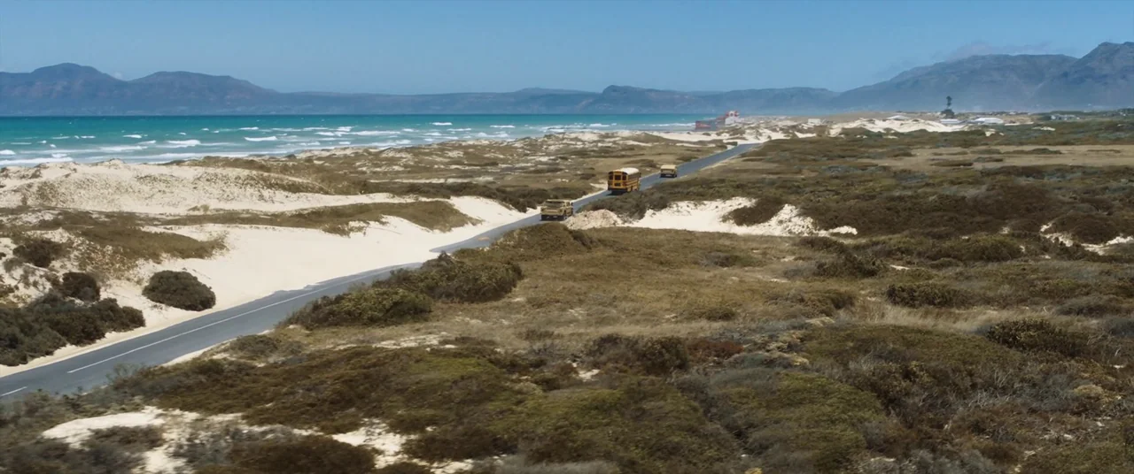 The Mauritanian (2021) movie still: establishing — A paved road snakes through grassy sand dunes alongside a turquoise ocean bay. Three vehicles…