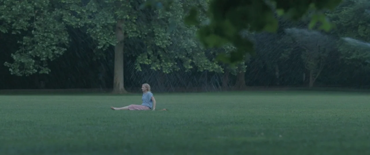 On Body and Soul (2017) movie still: wide — A vast, open green park features scattered trees and a light rain shower falling. A lone woman sits on…
