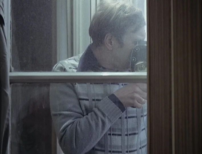Camera Buff (1979) movie still: medium — A man stands outside a window holding a small handheld camera to his eye. He is seen through the glass and…