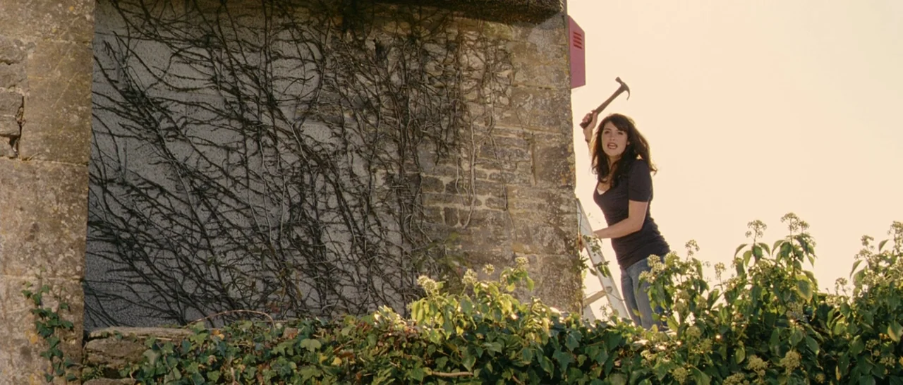 Tamara Drewe (2010) movie still: wide — A stone building wall is covered with climbing vines near a rural landscape. A woman stands on a ladder and…