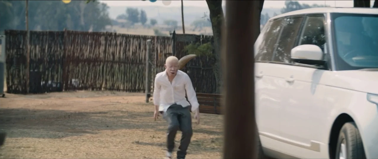 My Girlfriend's Father (2023) movie still: medium — An outdoor dirt driveway is enclosed by a rustic wooden stick fence. A man with albinism moves…