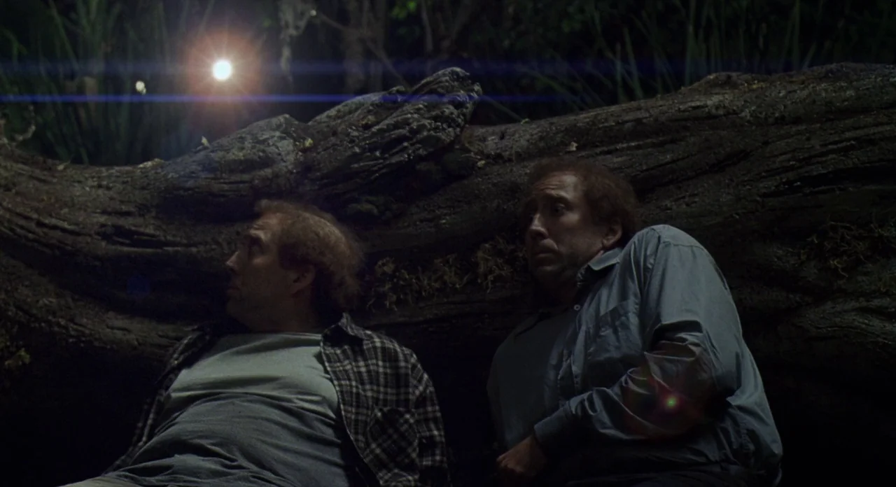 Adaptation (2002) movie still: medium — Two men sit against a large, fallen moss-covered tree log in a wooded area. They both look upward at a…