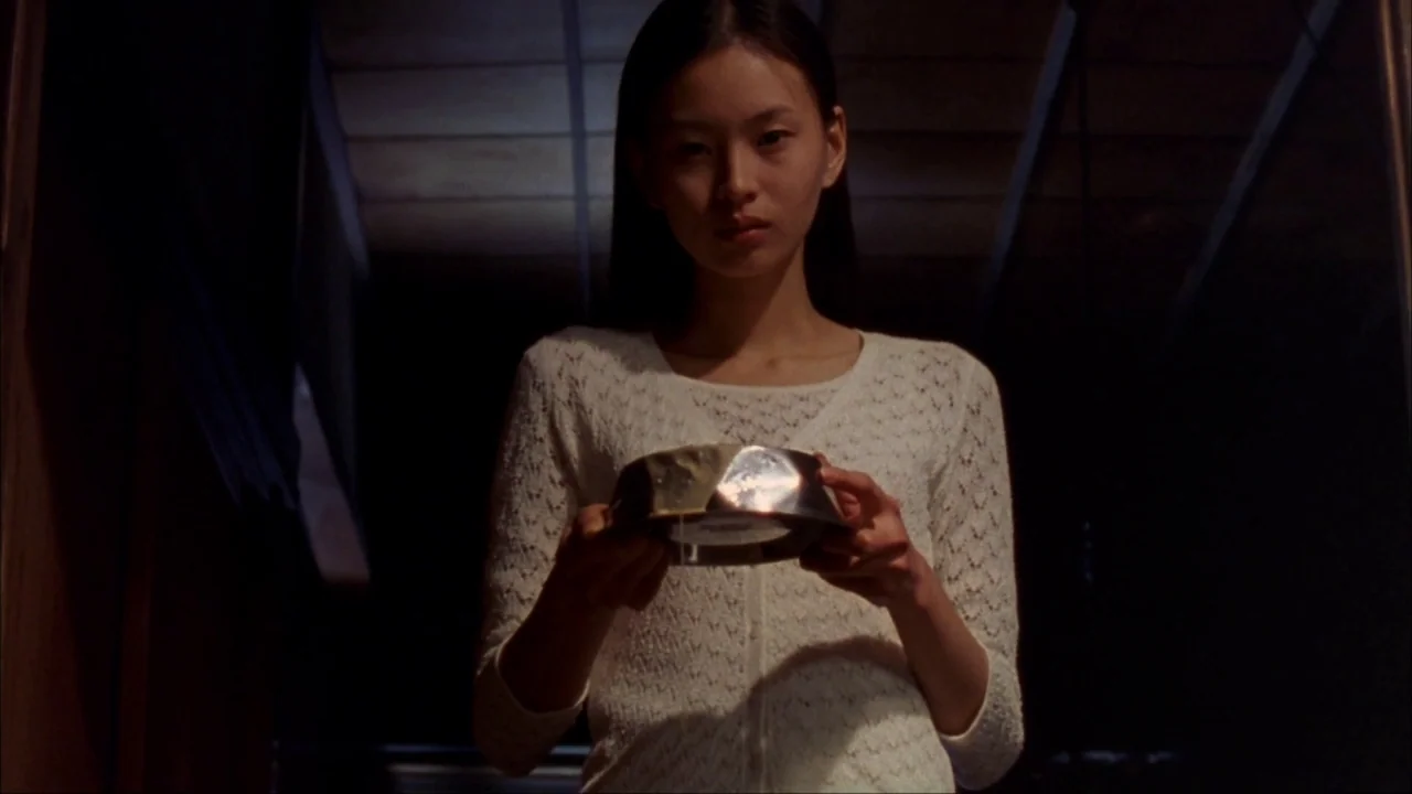 Audition (1999) movie still: medium — A young woman stands in a dimly lit, narrow corridor at night. She holds a small, reflective metal bowl…