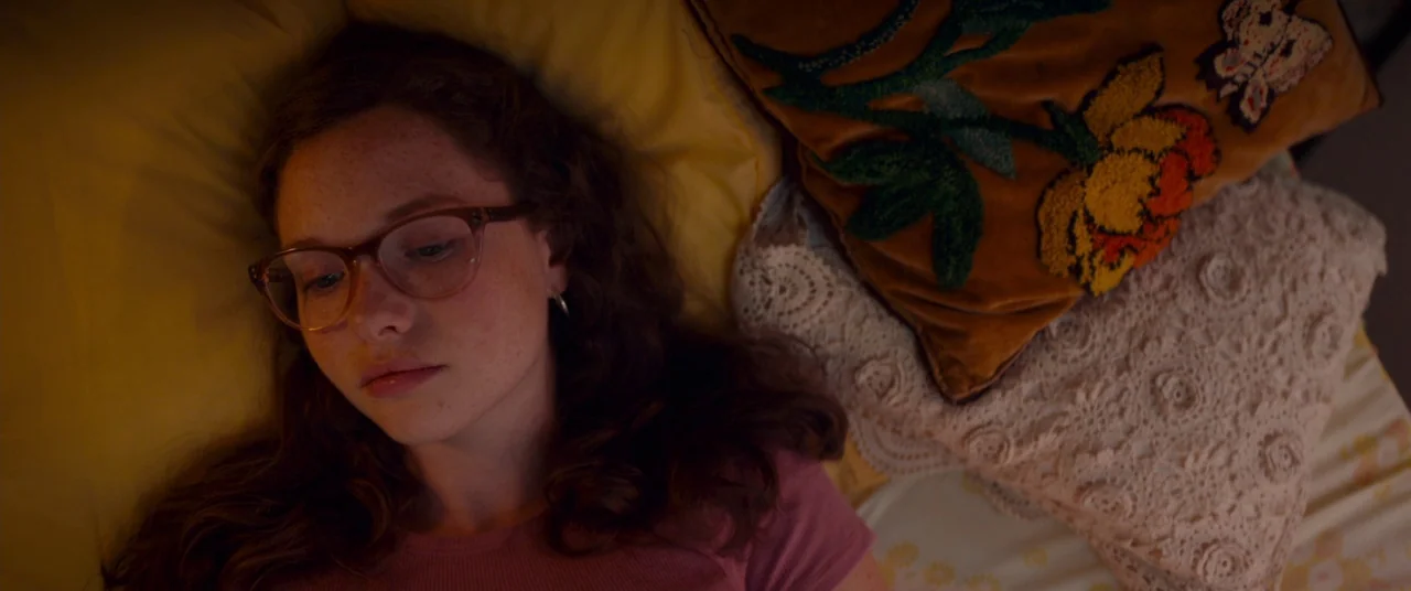 Scary Stories to Tell in the Dark (2019) movie still: closeup — A woman lies on a bed with yellow bedding and decorative embroidered pillows. She…