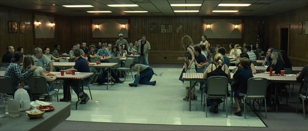 Take Shelter (2011) movie still: wide — A wood-paneled community hall features long tables with red checkered tablecloths and many seated people. A…