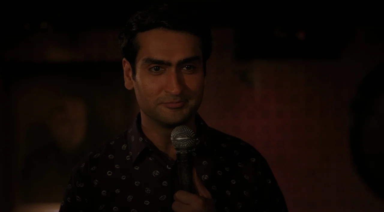 The Big Sick (2017) movie still: closeup — A dimly lit club environment with dark walls and subtle patterns. A man stands in front of a microphone…