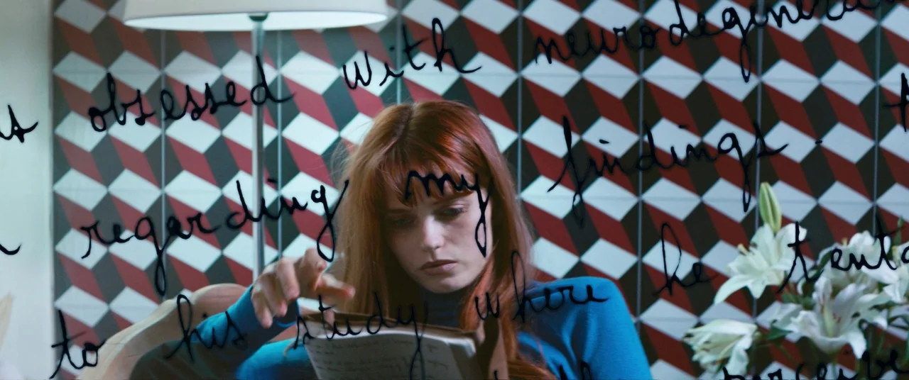 Elizabeth Harvest movie still: medium — A person sits in front of a wall with a repetitive red and white geometric pattern. Black cursive text is…