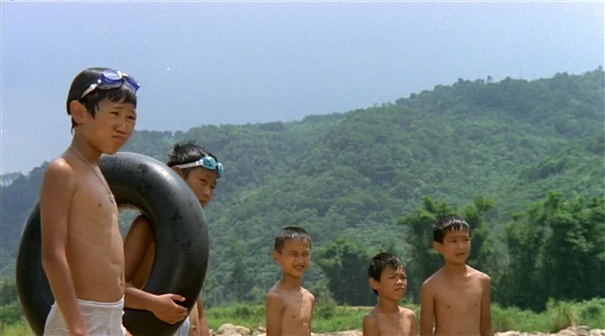 A Summer at Grandpa’s (1984) movie still: medium — A group of five young Asian boys stands on a rocky riverbank outdoors. The boys are mostly…