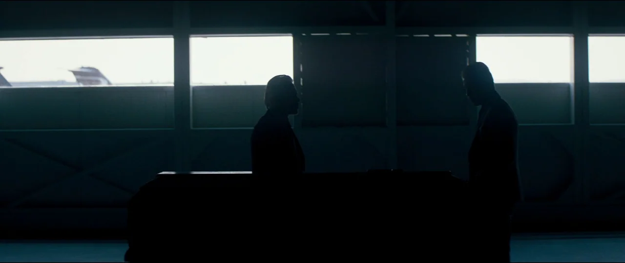 El Chicano (2018) movie still: wide — A dark, industrial structure features large horizontal windows overlooking an airfield. Two men in dark suits…