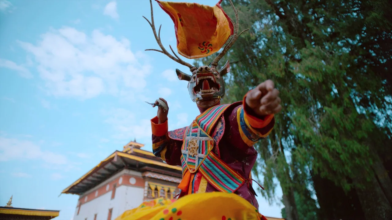 Awaken (2018) movie still: medium — A performer in a traditional Himalayan dance mask stands in a temple courtyard. The dancer points forward while…