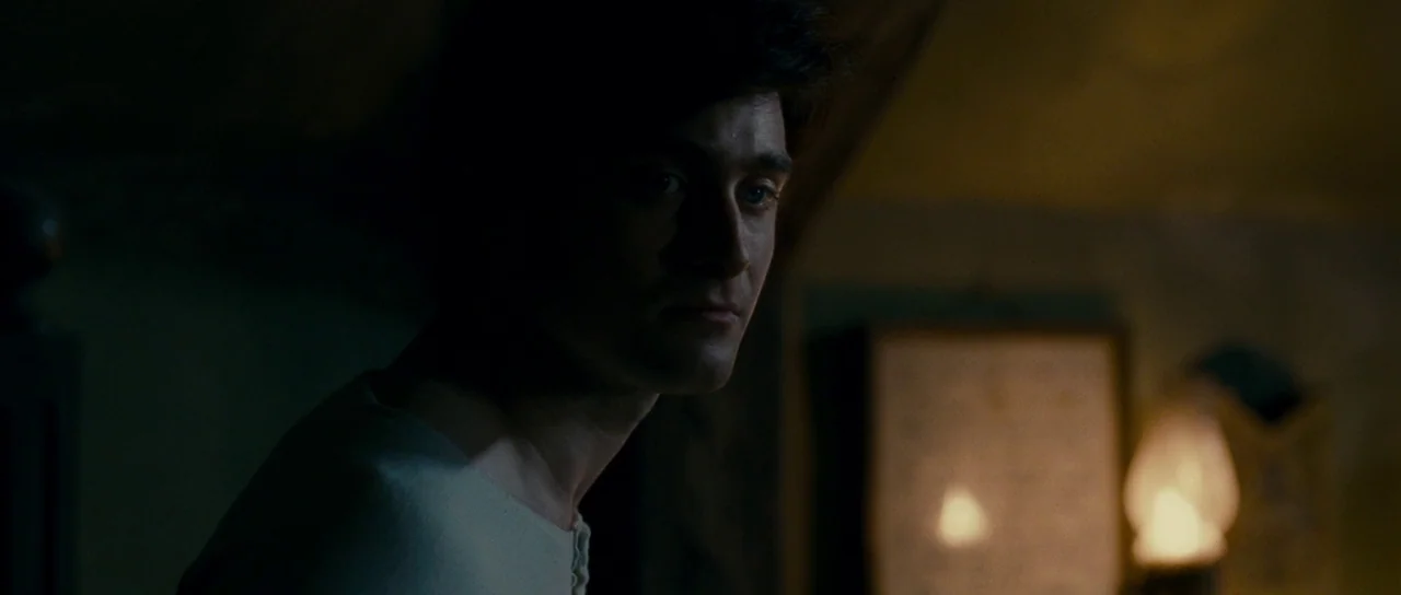The Woman in Black (2012) movie still: closeup — A dark indoor space at night with a blurred, glowing lamp in the background. A young man turns his…