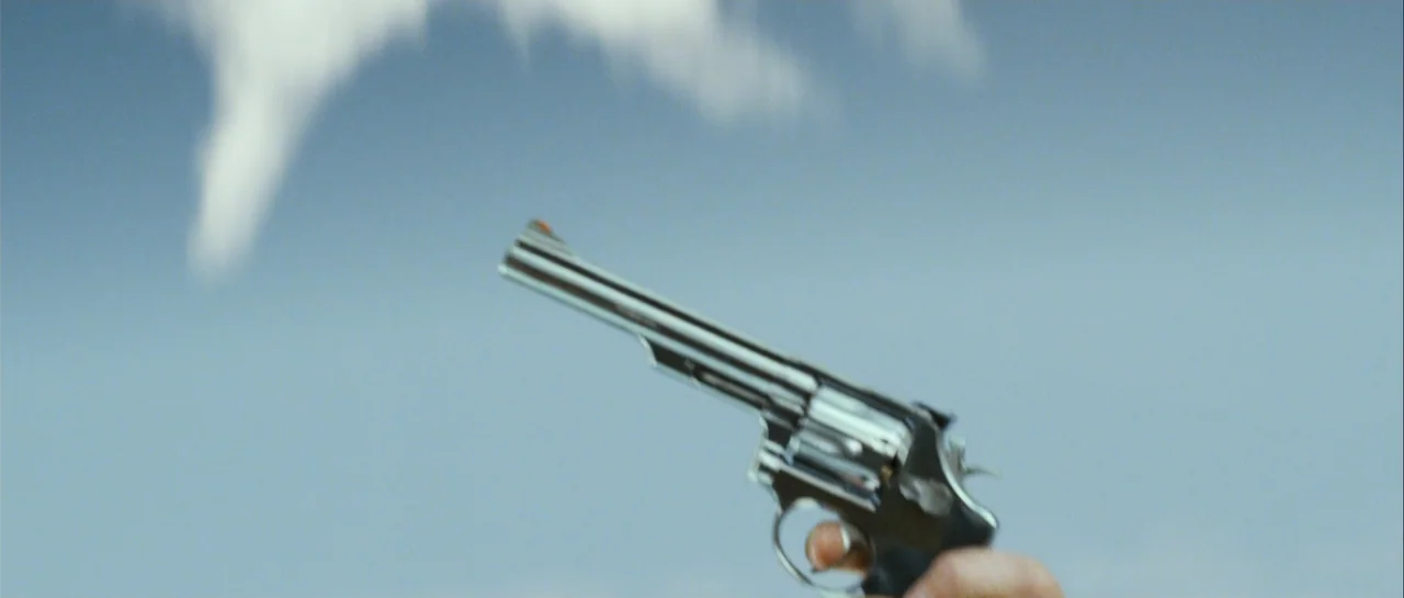 Shoot 'Em Up (2007) movie still: closeup — A close-up shot focuses on a metallic revolver held against a blue sky background. The hand holding the…