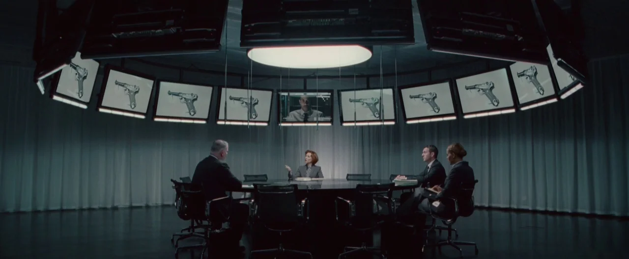 Hanna (2011) movie still: wide — A dark, modern circular conference room features white curtains lining the perimeter walls. Four individuals sit at…