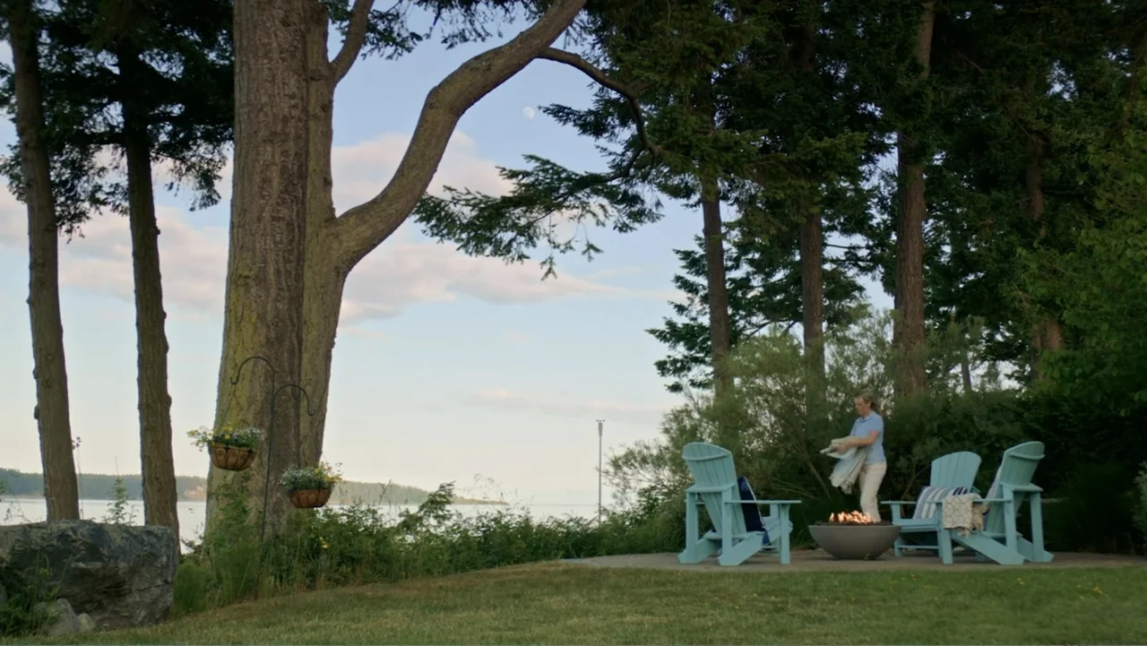 Making Waves (1998) movie still: wide — A stone patio sits alongside a lake with large evergreen trees and fire pit. A woman in a blue shirt stands…