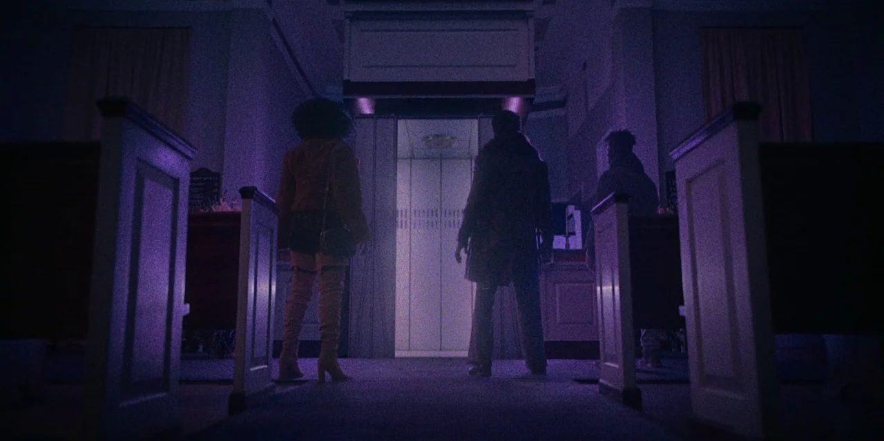 They Cloned Tyrone (2023) movie still: fullbody — A dimly lit interior hallway with white partitions leading to an illuminated elevator door. Two…