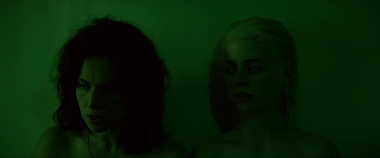Ema (2019) movie still: medium — A plain wall serves as the backdrop for two women in a dark space. They stand together under a strong green light…