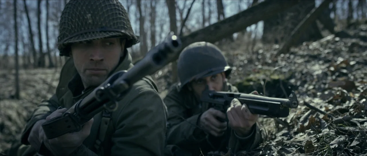 Battle of the Bulge (1965) movie still: medium — A dense woodland environment with fallen leaves covering the ground and blurred trees behind. Two…