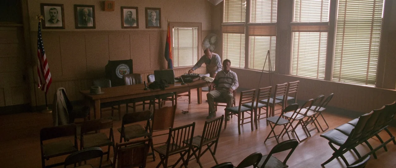 Fire In The Sky (1993) movie still: wide — A quiet hall has rows of folding chairs facing a long wooden front table. Two men perform an audio…