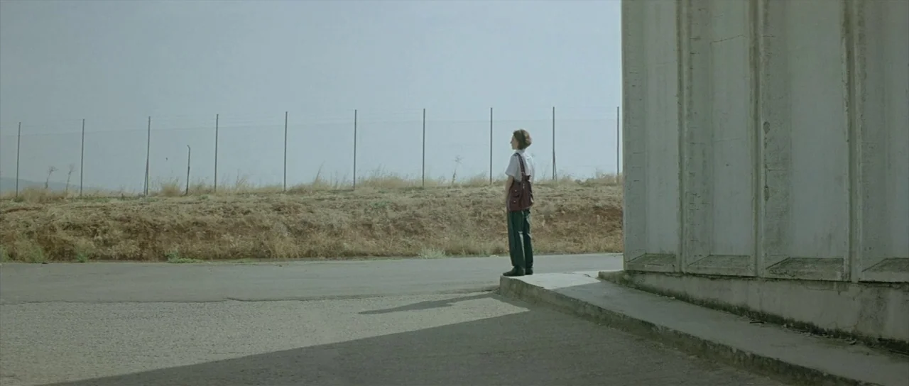 Dogtooth movie still: wide — A desolate paved area with a concrete wall and a wire fence. A young woman stands alone on the curb, looking out at the…