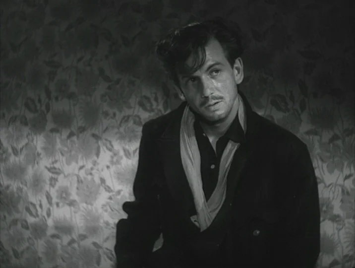 Crisis (1946) movie still: medium — A man stands indoors against a backdrop of heavily patterned Victorian-style floral wallpaper. The subject wears…
