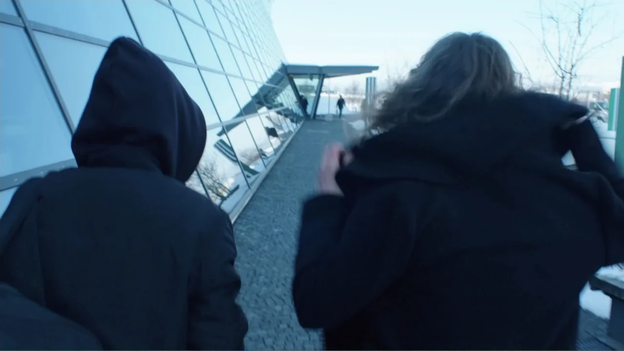 Code of Trust (2019) movie still: medium — A modern glass building exterior with a paved path runs alongside a wall. Two figures in dark clothing…