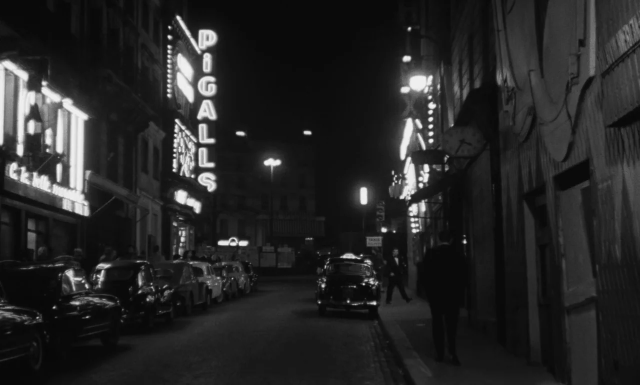 Witness In The City (1959) movie still: wide — A night view of a Paris street featuring a vertical neon sign reading Pigalle. Several vintage cars…