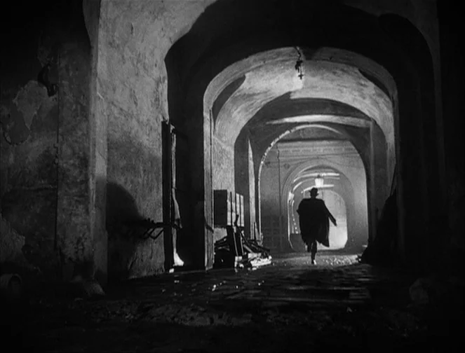 The Third Man (1949) movie still: wide — An old stone passageway with vaulted archways leads into a misty night setting. A lone man in a trench coat…
