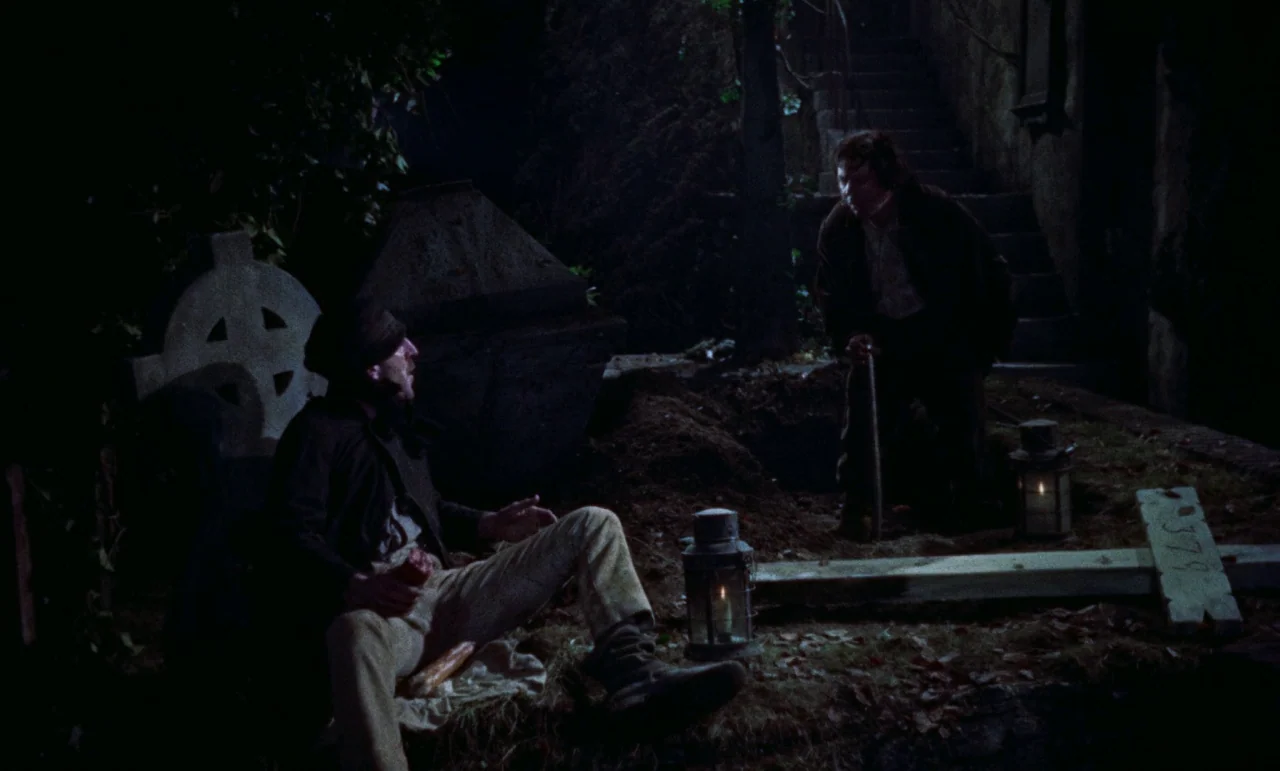 The Revenge of Frankenstein movie still: wide — A dark cemetery at night features an open grave and scattered wooden grave markers. One man sits by…