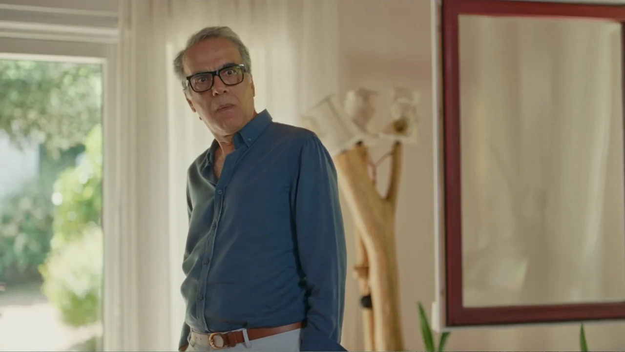 Paranormal Cuma (2024) movie still: medium — A sunlit room features sheer white curtains and a minimalist wooden lamp structure. An older man stands…