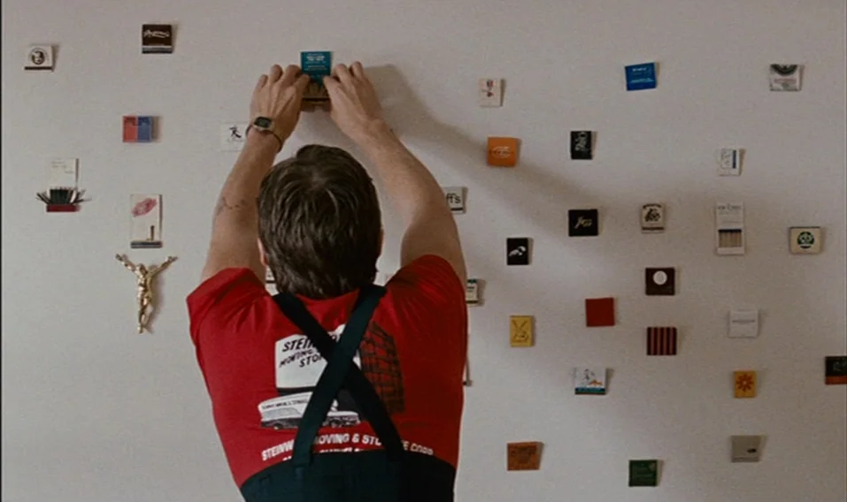 Blue Valentine (2010) movie still: medium — A man stands before a blank, off-white wall decorated with various small matchbook covers. He reaches up…