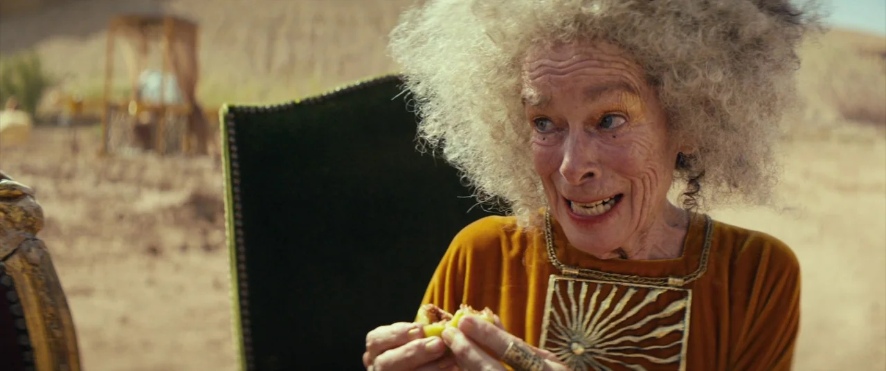 Seneca: On The Creation Of Earthquakes (2023) movie still: medium — An elderly woman sits in a velvet chair amidst a vast arid desert landscape. She…