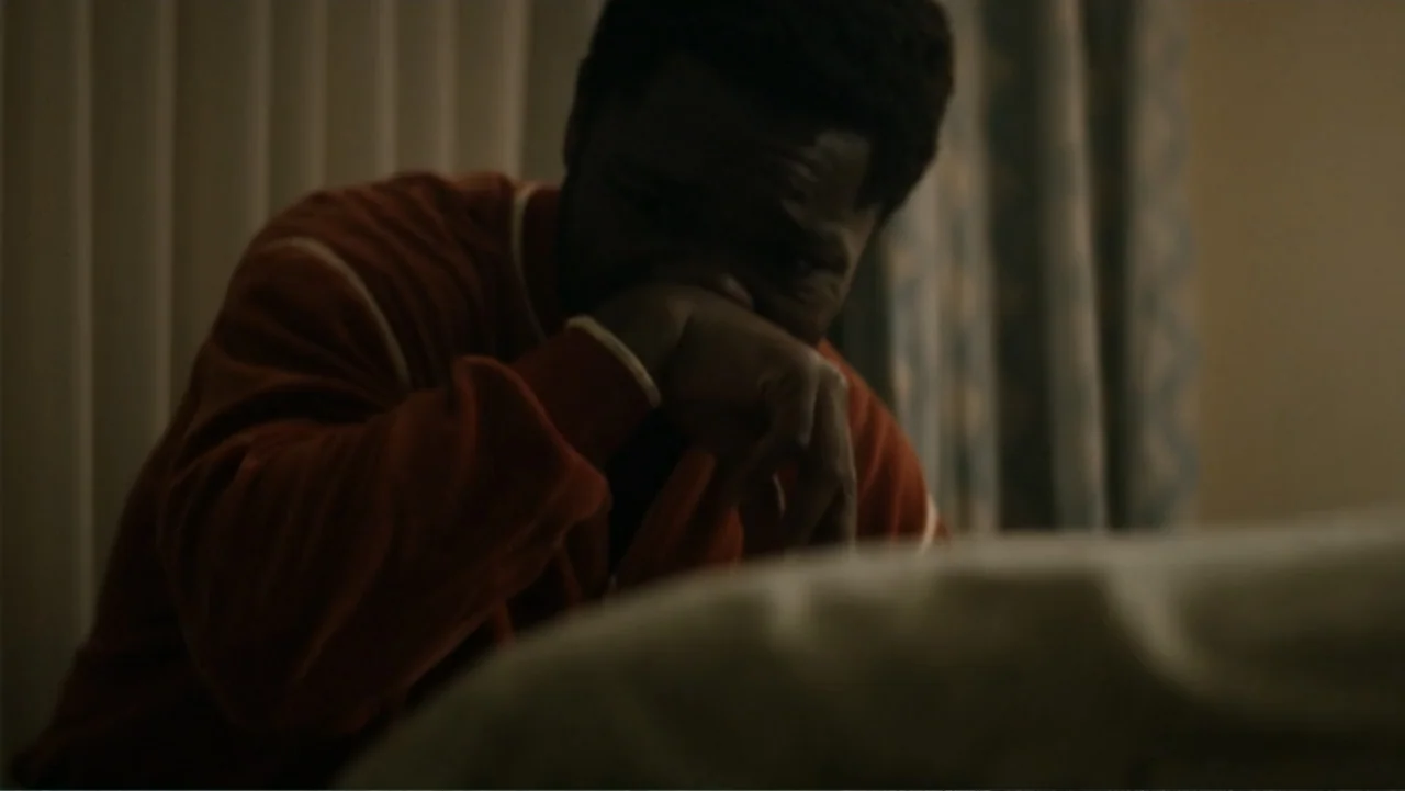 Nightingale (2014) movie still: medium — A dimly lit bedroom features vertical blinds and a bed in the foreground. A man wearing a red shirt sits on…