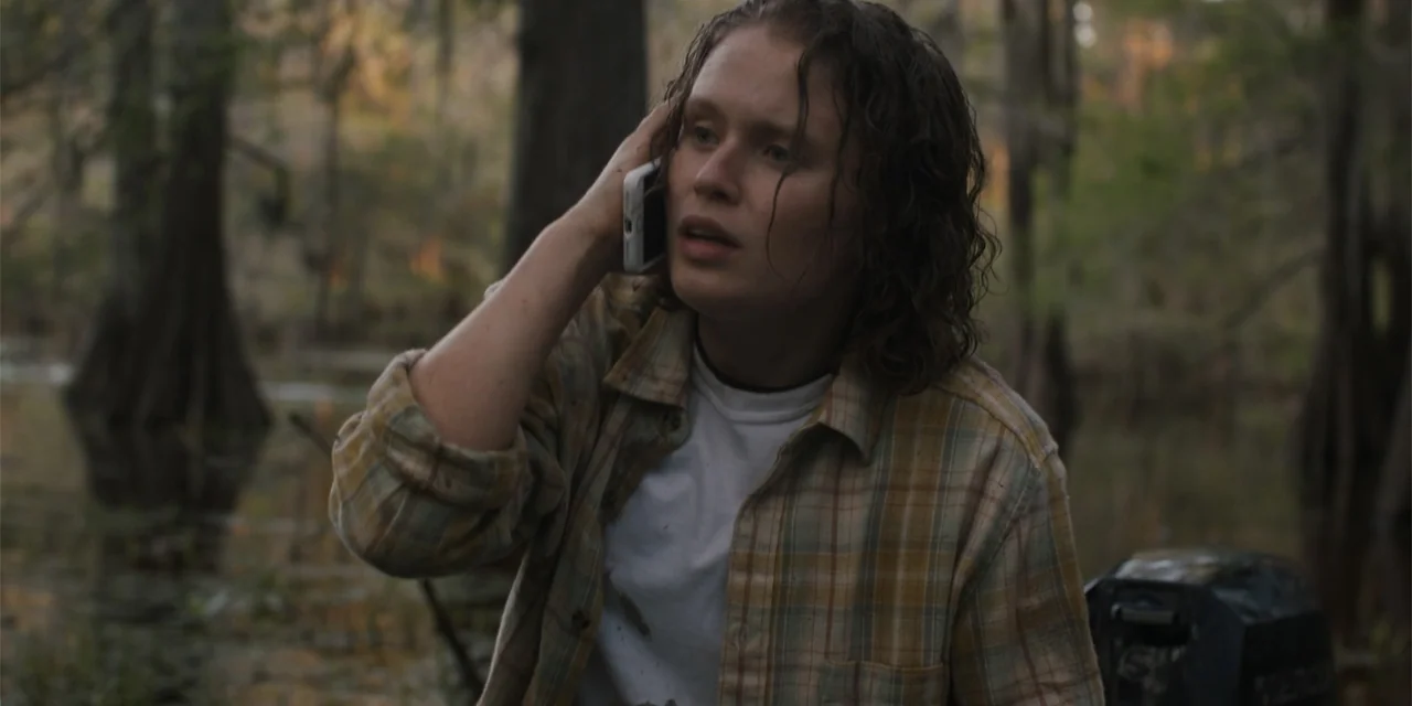 Caddo Lake (2024) movie still: medium — A dark swamp filled with moss-covered cypress trees at dusk. A woman in a plaid shirt speaks on a smartphone…