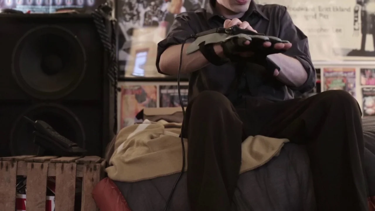 Buzzard (2014) movie still: medium — A cluttered room contains a bed covered by a light-colored blanket and large speakers. A person sitting on the…