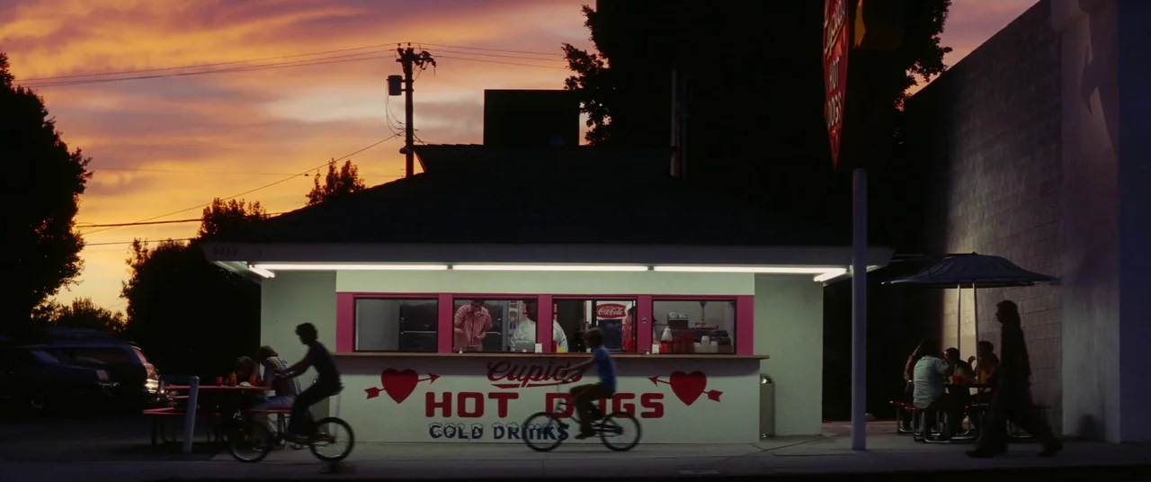 Licorice Pizza (2021) movie still: wide — A small stand-alone fast food restaurant sits under a sunset sky with orange clouds. Several people ride…