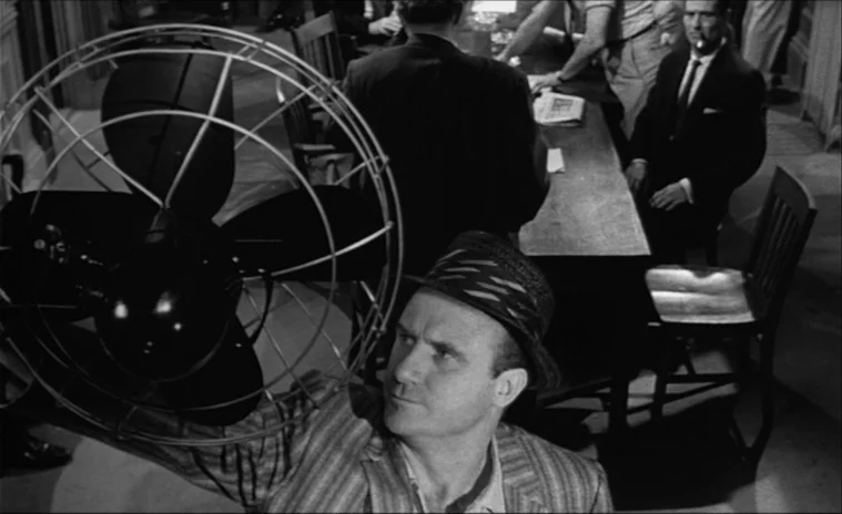 12 Angry Men (1957) movie still: medium — An office room features a large, dark pedestal fan in the foreground. A man looks up at the fan while…
