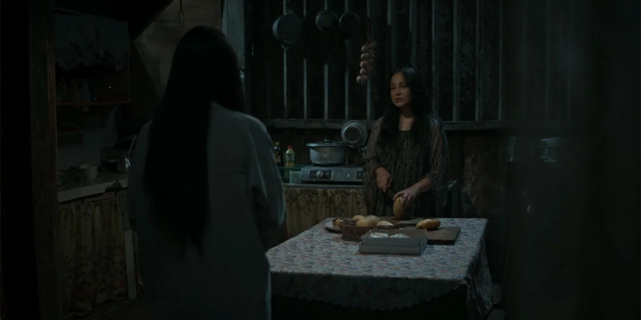 Nokturno (2024) movie still: medium — A dimly lit room with wood-paneled walls and a small covered table. A woman stands in the center slicing fruit…