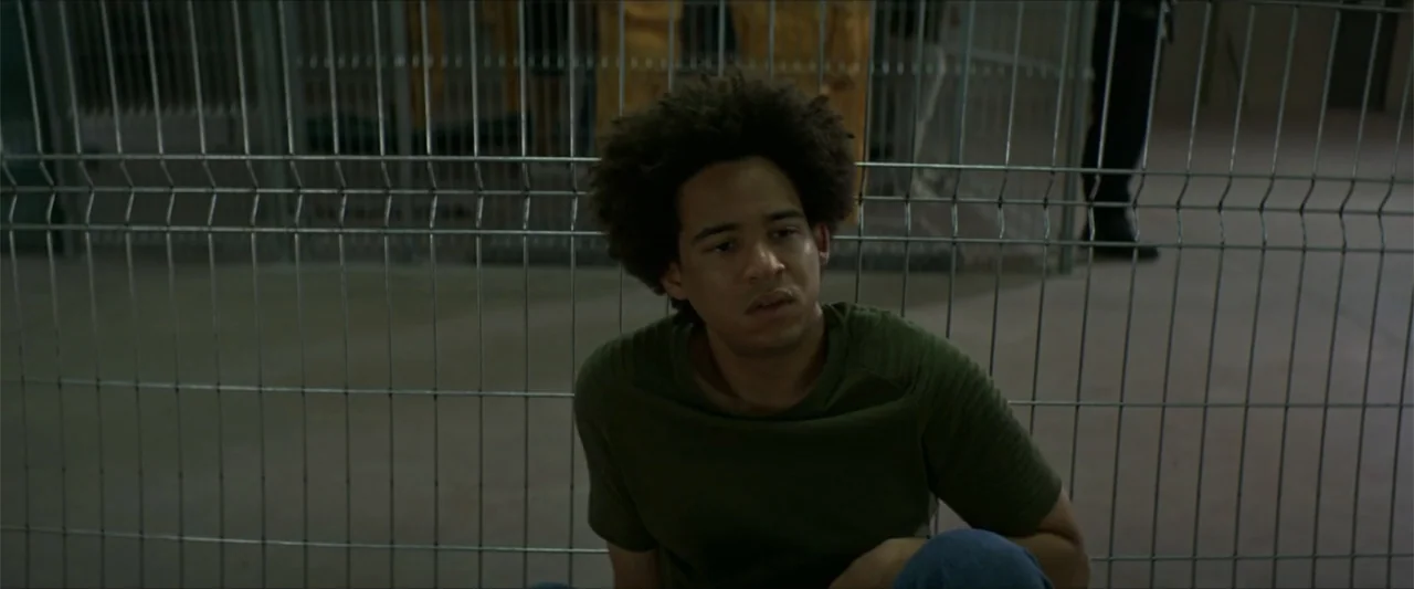 American Carnage (2022) movie still: medium — A dimly lit interior space features a metal wire security fence in the foreground. A young man with…