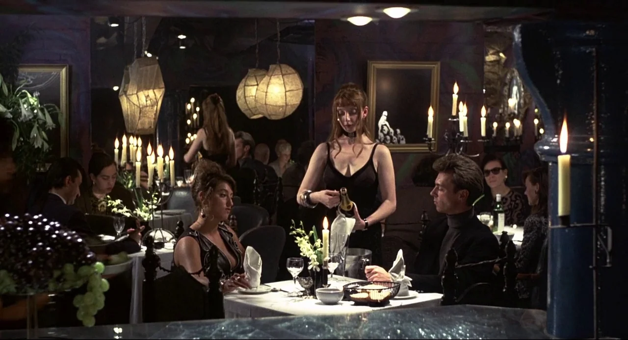 Naked (1993) movie still: wide — A dimly lit upscale restaurant features dark purple walls and numerous lit candle centerpieces. A waitress in a…