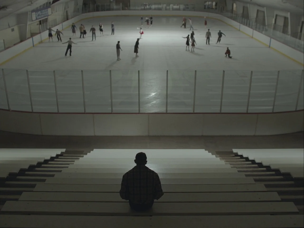 The Mountain (2018) movie still: wide — An empty ice rink seen from the top of the tiered bleacher seating. A lone figure sits in the middle of the…