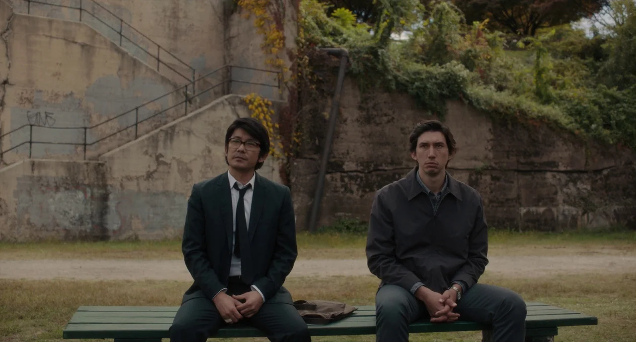Paterson (2016) movie still: fullbody — A public park area features a weathered green bench in front of a concrete staircase. Two men in…